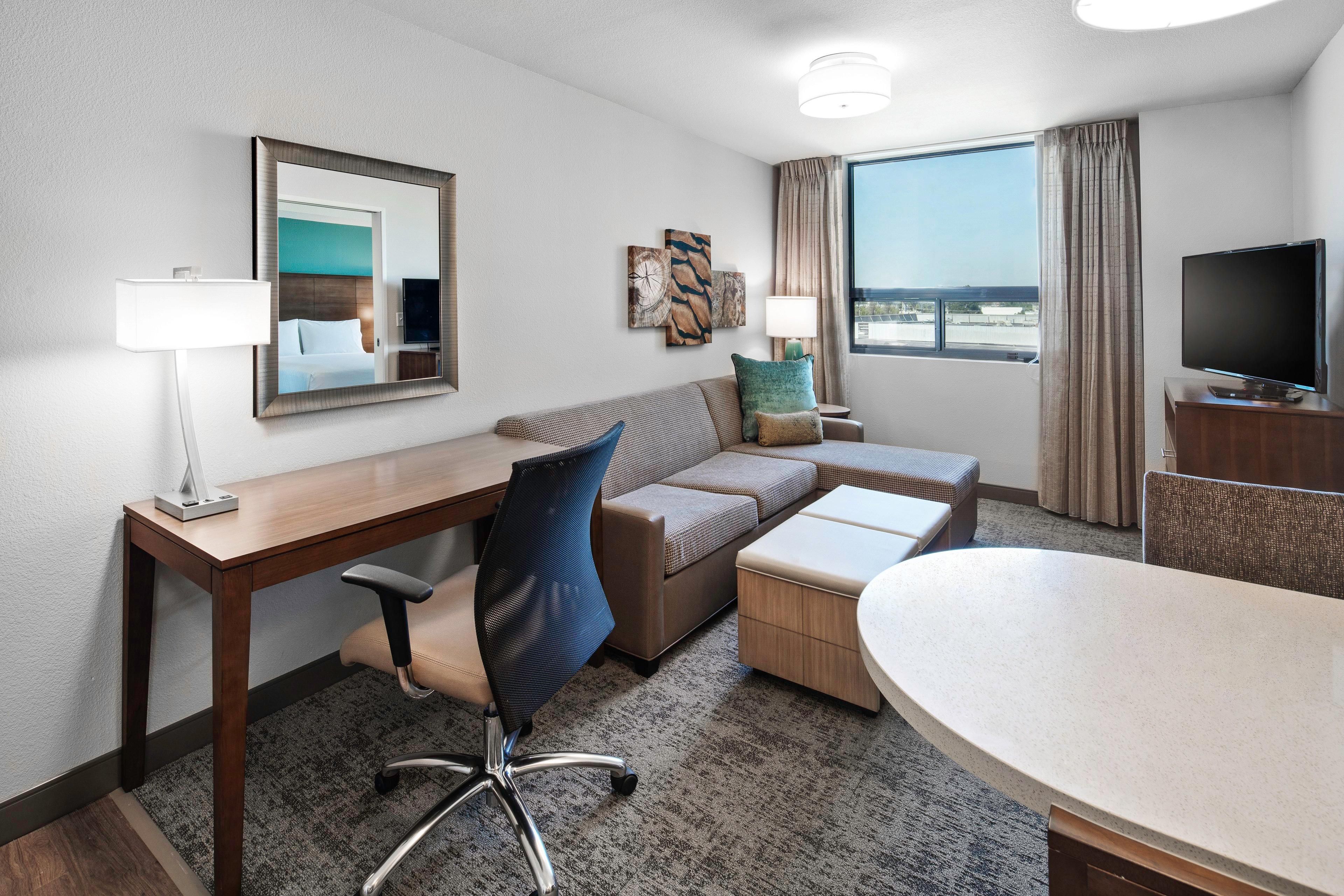 A hotel room with a desk, a sofa, a TV, and a large window.