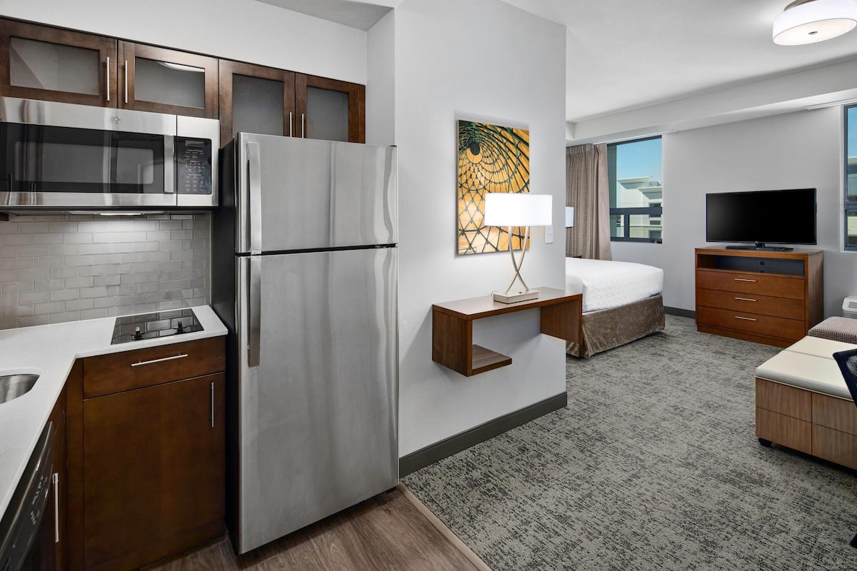 A modern hotel room with a kitchenette, a bed, a TV, and a view of the city.