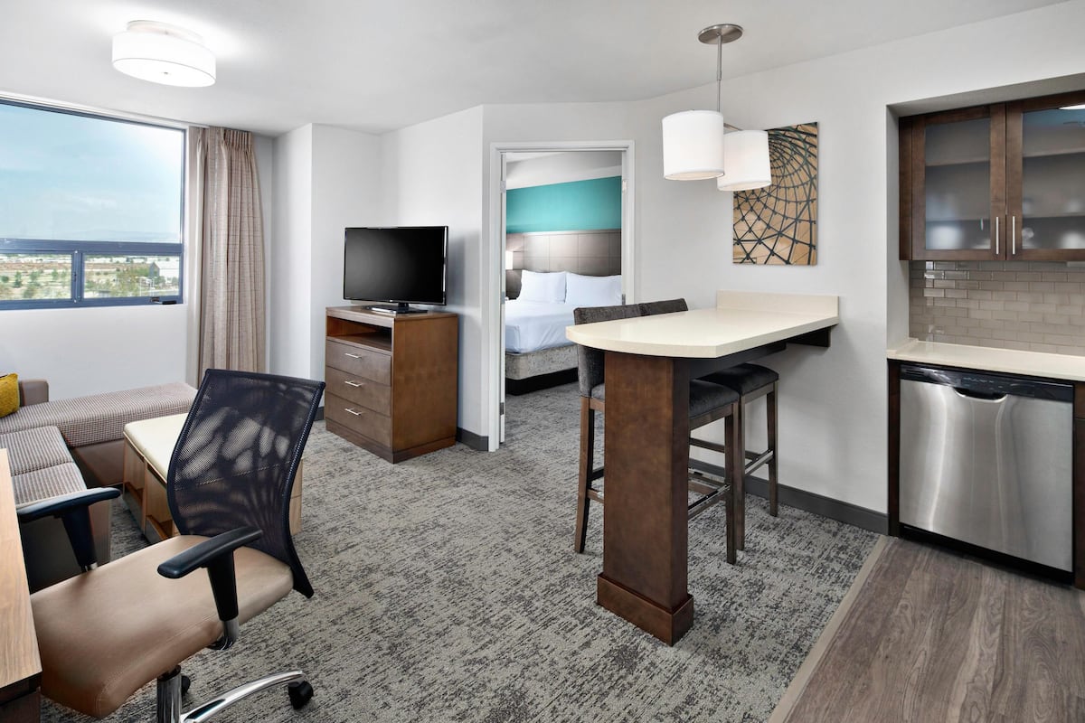 A hotel room with a bed, a TV, a desk with a chair, a dining area, and a kitchen.