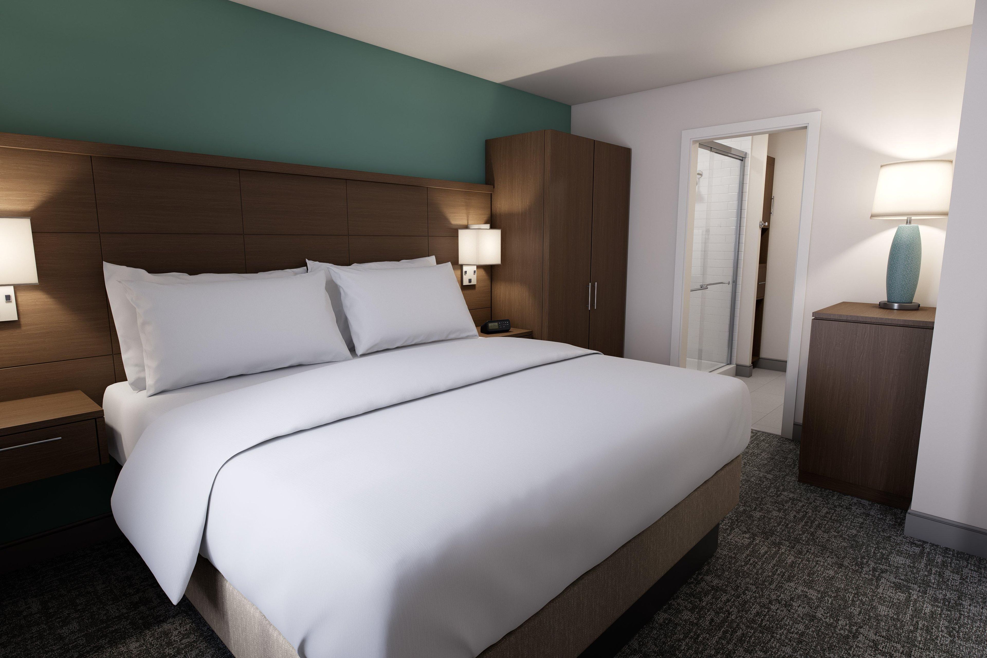 A hotel room with a large bed, wooden headboard, two bedside lamps, a nightstand, a wardrobe, and a door leading to a bathroom.