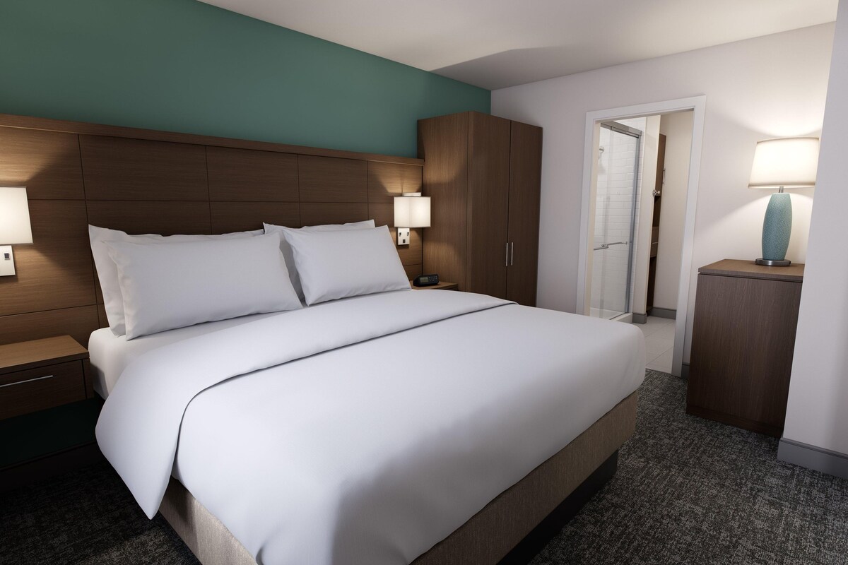 Una habitación de hotel con una cama grande, cabecera de madera, dos lámparas de noche, una mesita de noche, un armario y una puerta que da a un baño.