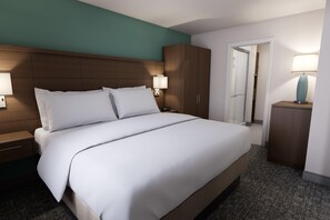 Una habitación de hotel con una cama grande, cabecera de madera, dos lámparas de noche, una mesita de noche, un armario y una puerta que da a un baño.
