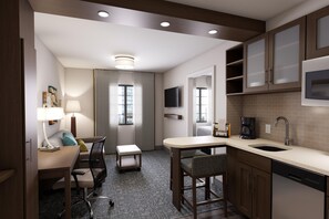 A compact hotel room with a kitchenette, a desk, a chair, a small table, and a TV mounted on the wall. - Staybridge Suites Irvine - John Wayne Airport IHG (Costa Mesa)