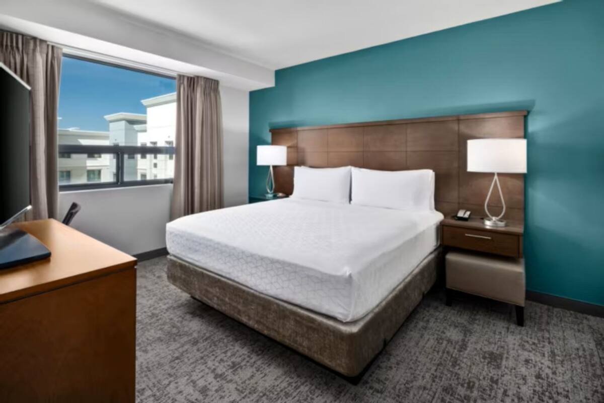 A hotel room with a large bed, a desk, a chair, a nightstand with a lamp, and a view of buildings through the window.