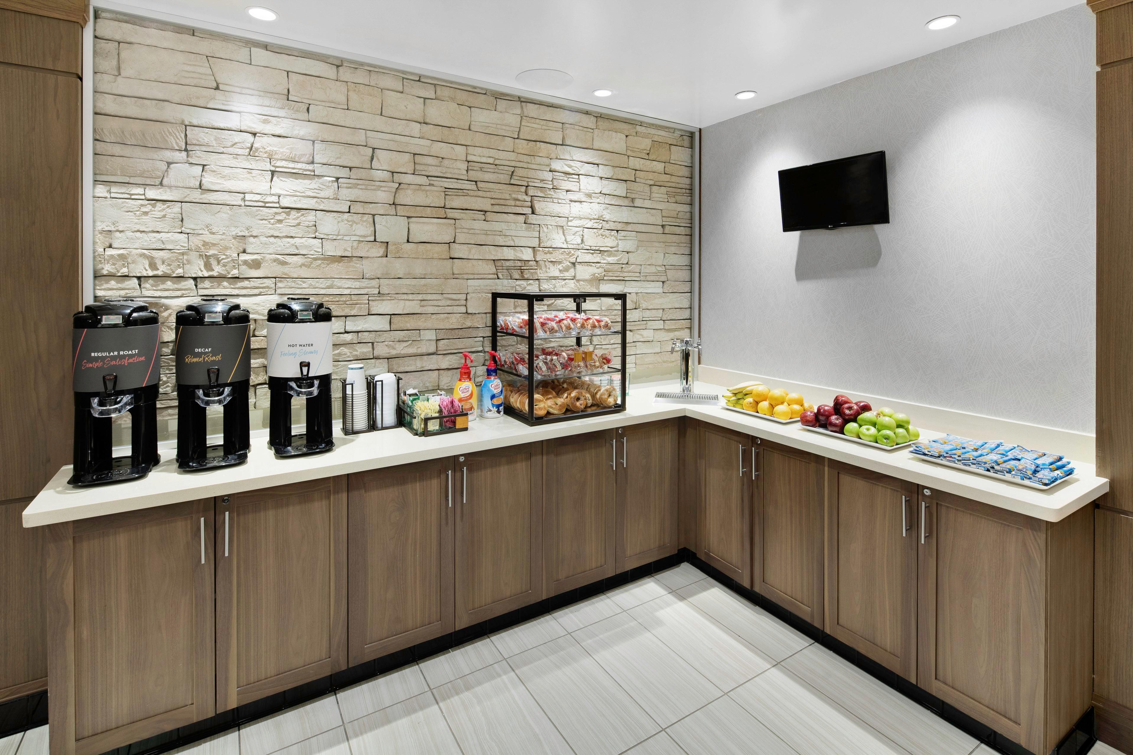 A modern kitchen with a stone backsplash, coffee makers, a fruit display, and a flat-screen TV.