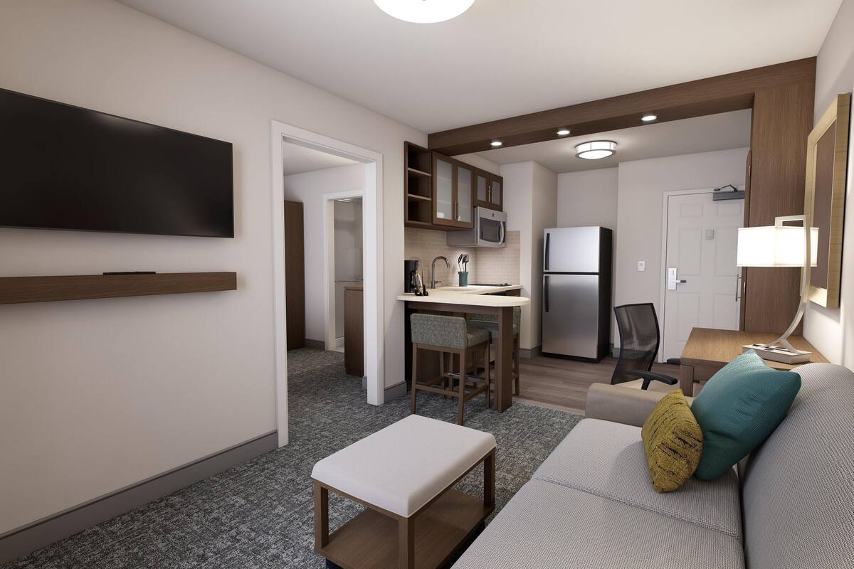A modern living room with a flat-screen TV, a sofa with cushions, a small table, and a kitchen area with a microwave, refrigerator, and dining table.