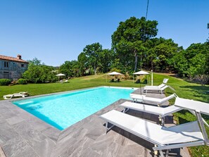 Une piscine entourée d’une pelouse et d’arbres, avec des chaises longues et des parasols à proximité.