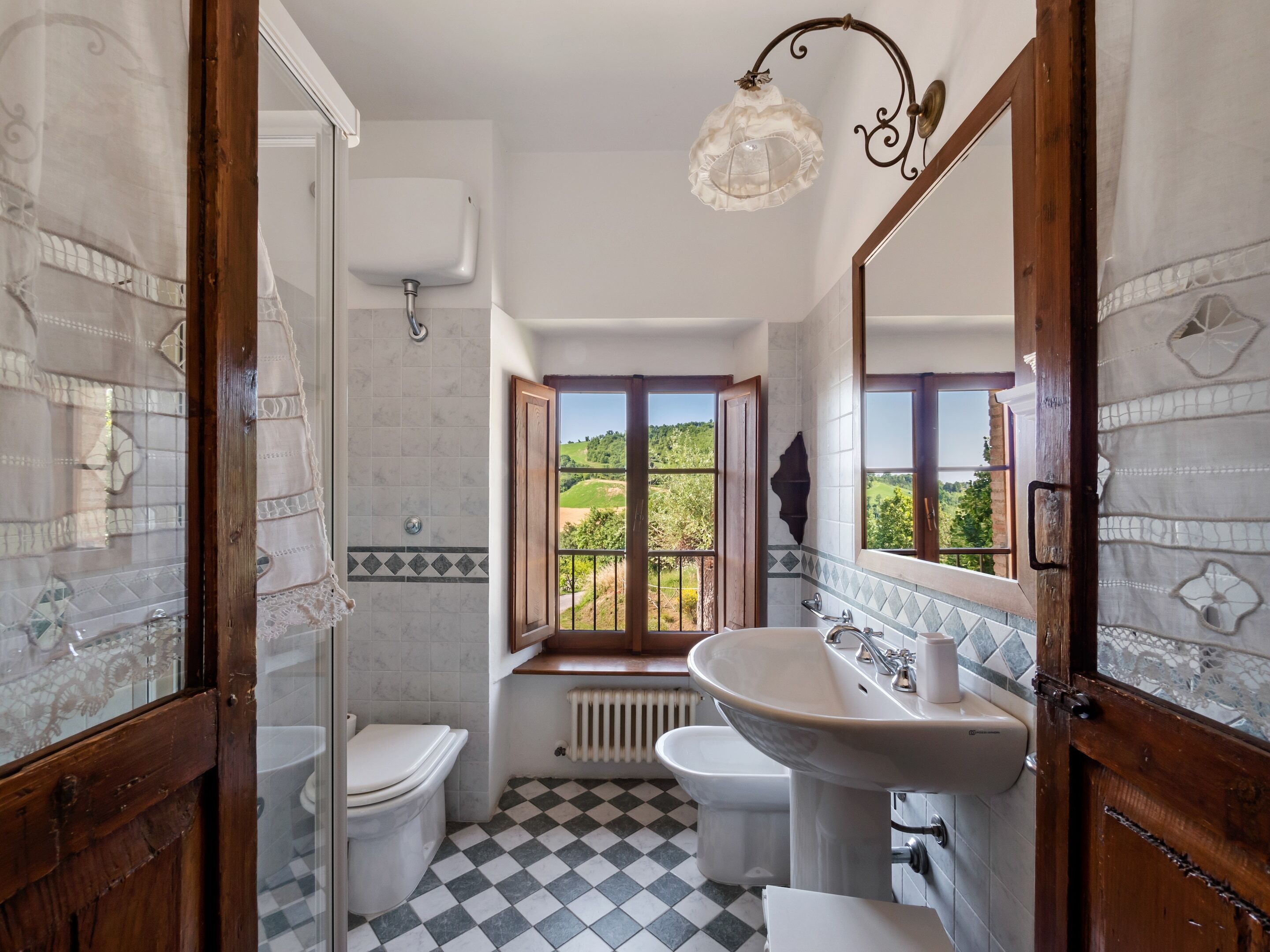 A bathroom with a checkered floor, a large window with a view of greenery, a white sink, and a toilet.