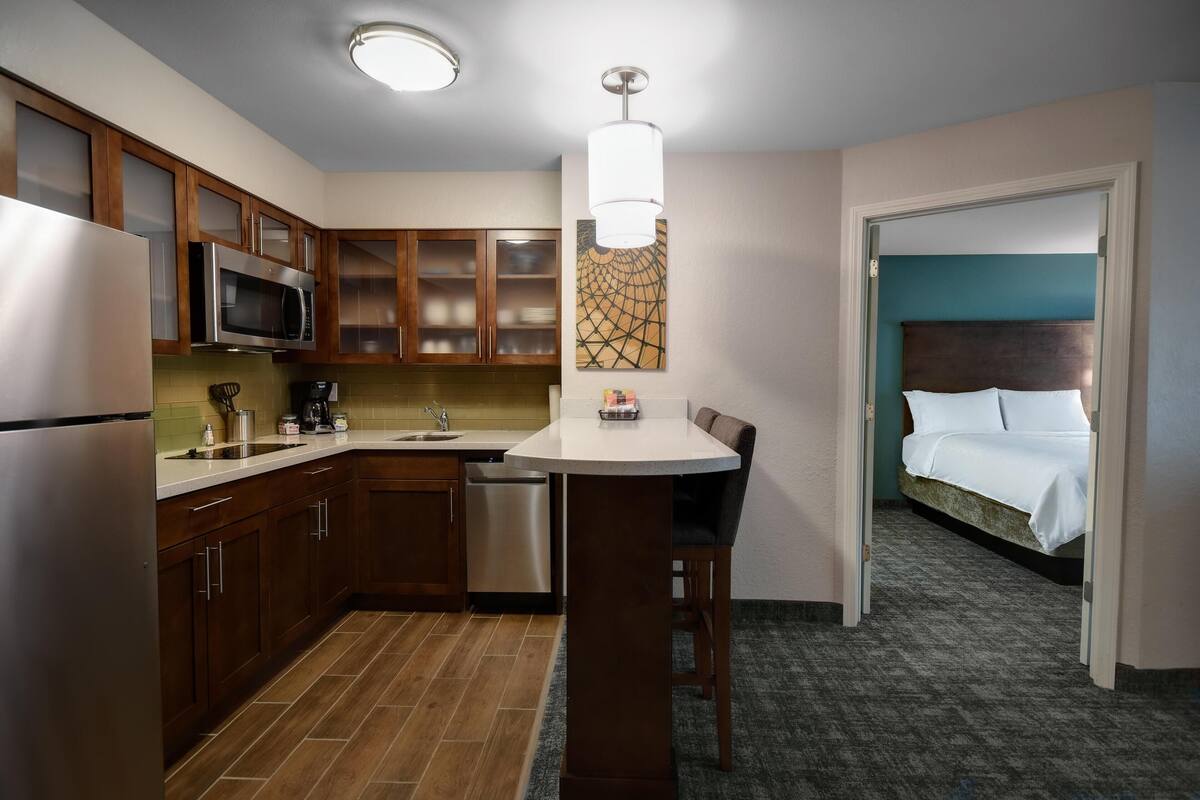 A compact hotel room with a kitchenette, a bed with a headboard, and a small dining area.