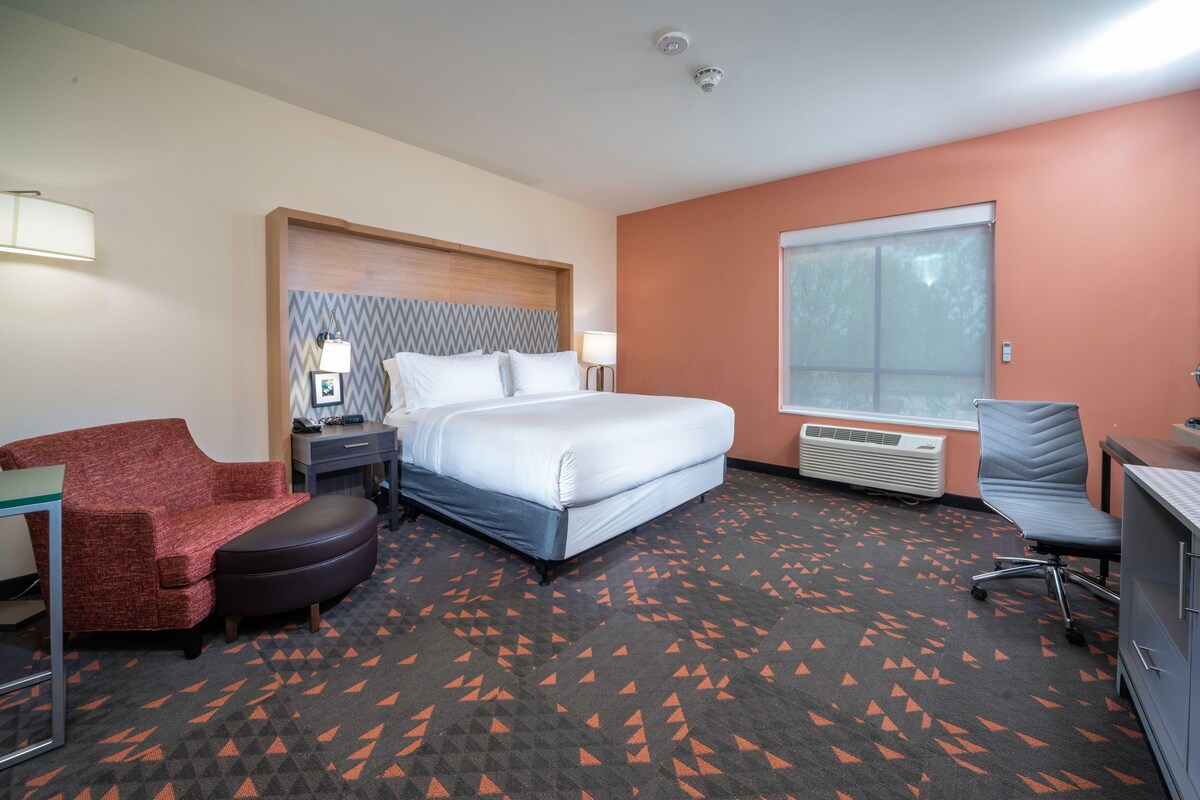 A hotel room with a bed, a red armchair, a desk, and a large window.