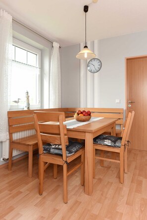 Un coin repas avec une table et des chaises en bois, un saladier de fruits, une horloge et une fenêtre avec des rideaux.