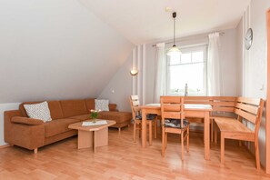 Un salon chaleureux avec un canapé marron, une table à manger en bois et des chaises. On y trouve une fenêtre avec des rideaux, une horloge au mur et un luminaire au plafond.