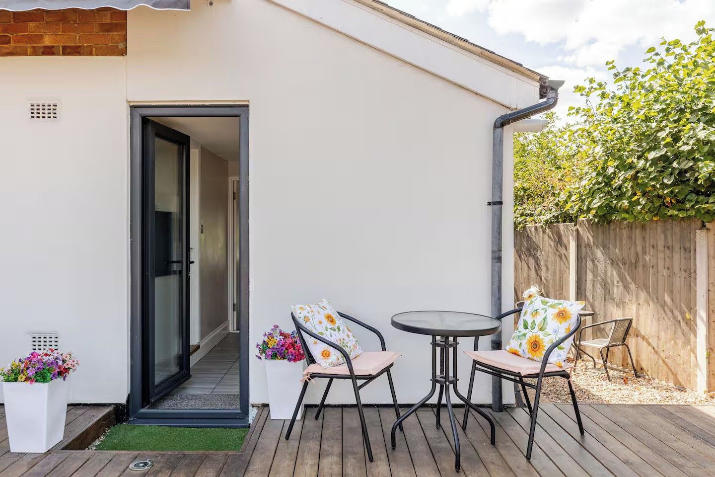 A patio with two chairs and a table, a wooden deck, a white wall, and a wooden fence.