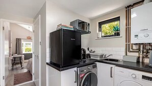 Une cuisine compacte équipée d’un lave-linge Bosch, d’un réfrigérateur et d’une fenêtre donnant sur un espace verdoyant.