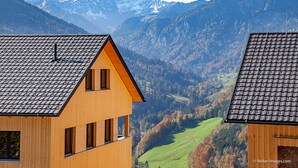 A wooden house with a sloped roof, multiple windows, and a view of a valley with mountains in the background.
