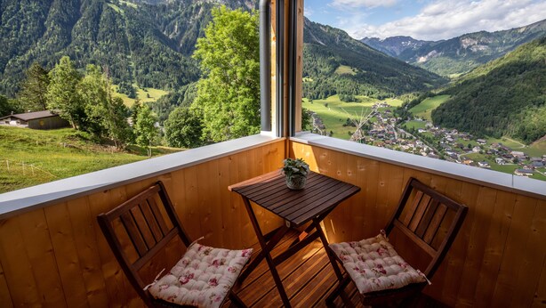 A wooden balcony with a table and two chairs, offering a view of a mountainous landscape.