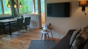 A modern living room with a dark sofa, a flat-screen TV mounted on the wall, a dining area with a wooden table and chairs, and large windows offering a view of greenery.