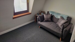 Une petite pièce accueillante avec un plafond en pente, une fenêtre avec des stores, un canapé gris agrémenté de coussins à carreaux et une petite table d’appoint sur laquelle est posé un objet décoratif.