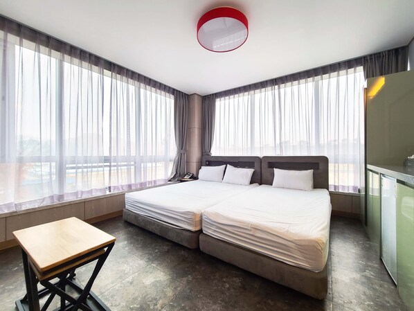 A hotel room with a double bed, a small wooden table, and a large window with sheer curtains.