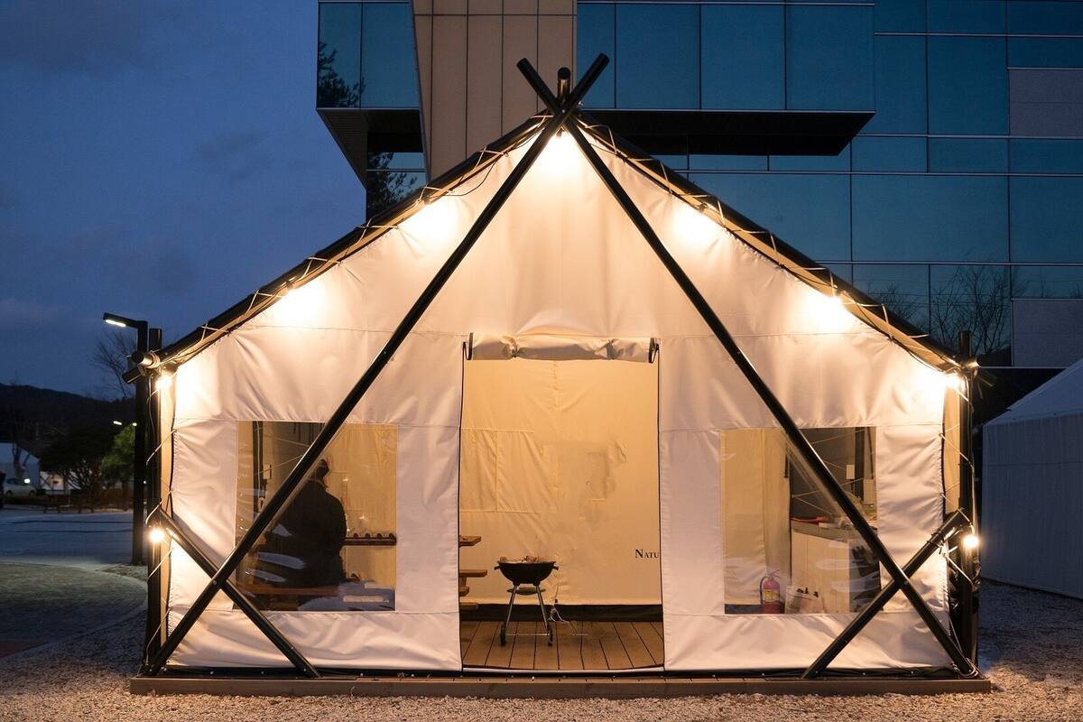 Una estructura tipo tienda de campaña con una zona de cocina central, ubicada frente a un edificio moderno al anochecer.