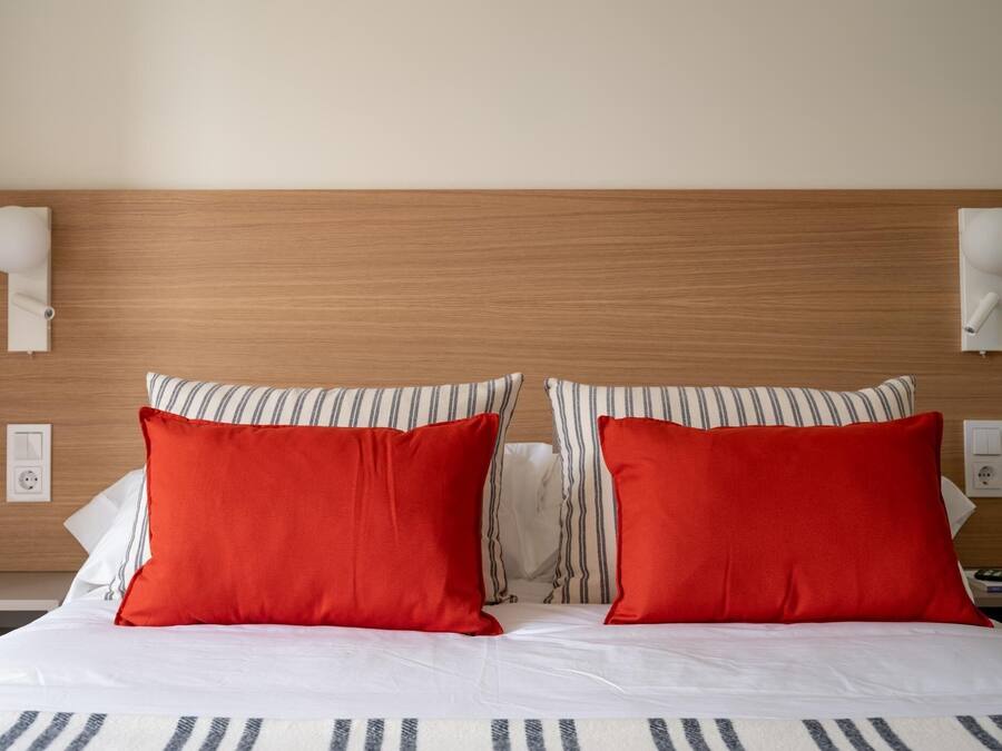 Habitación de hotel con cabecera de madera, dos almohadas rojas y almohadas a rayas, además de apliques de pared blancos.