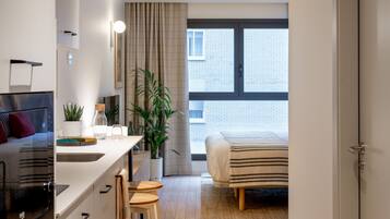 Una cucina moderna con isola centrale, zona pranzo con sgabelli, un letto con copriletto a righe e un'ampia finestra vista su un edificio in mattoni.
