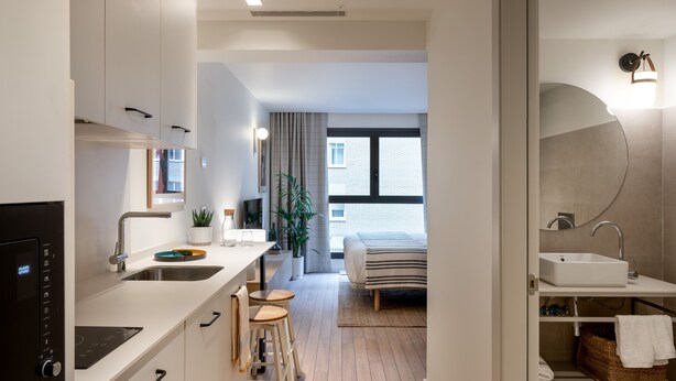 A modern kitchen with a built-in oven, a dining area with stools, a bed, and a bathroom with a sink and mirror. - Aparthotel Libere Vitoria (Vitoria-Gasteiz)