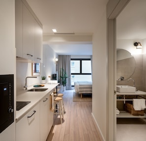 A modern kitchen with a built-in oven, a dining area with stools, a bed, and a bathroom with a sink and mirror. - Aparthotel Libere Vitoria (Vitoria-Gasteiz)