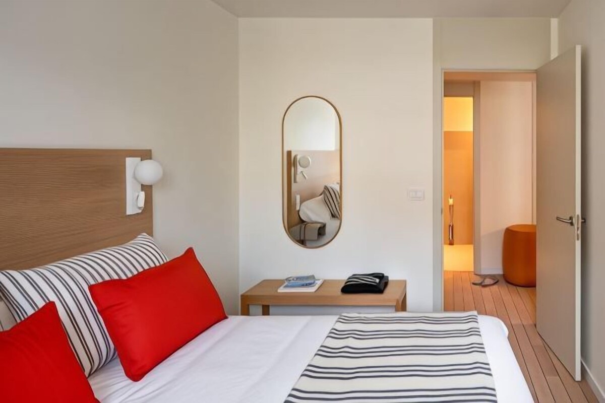 Ein Hotelzimmer mit einem Bett, einem Nachttisch mit Buch und Telefon, einem Spiegel und einer Tür zu einem weiteren Raum.