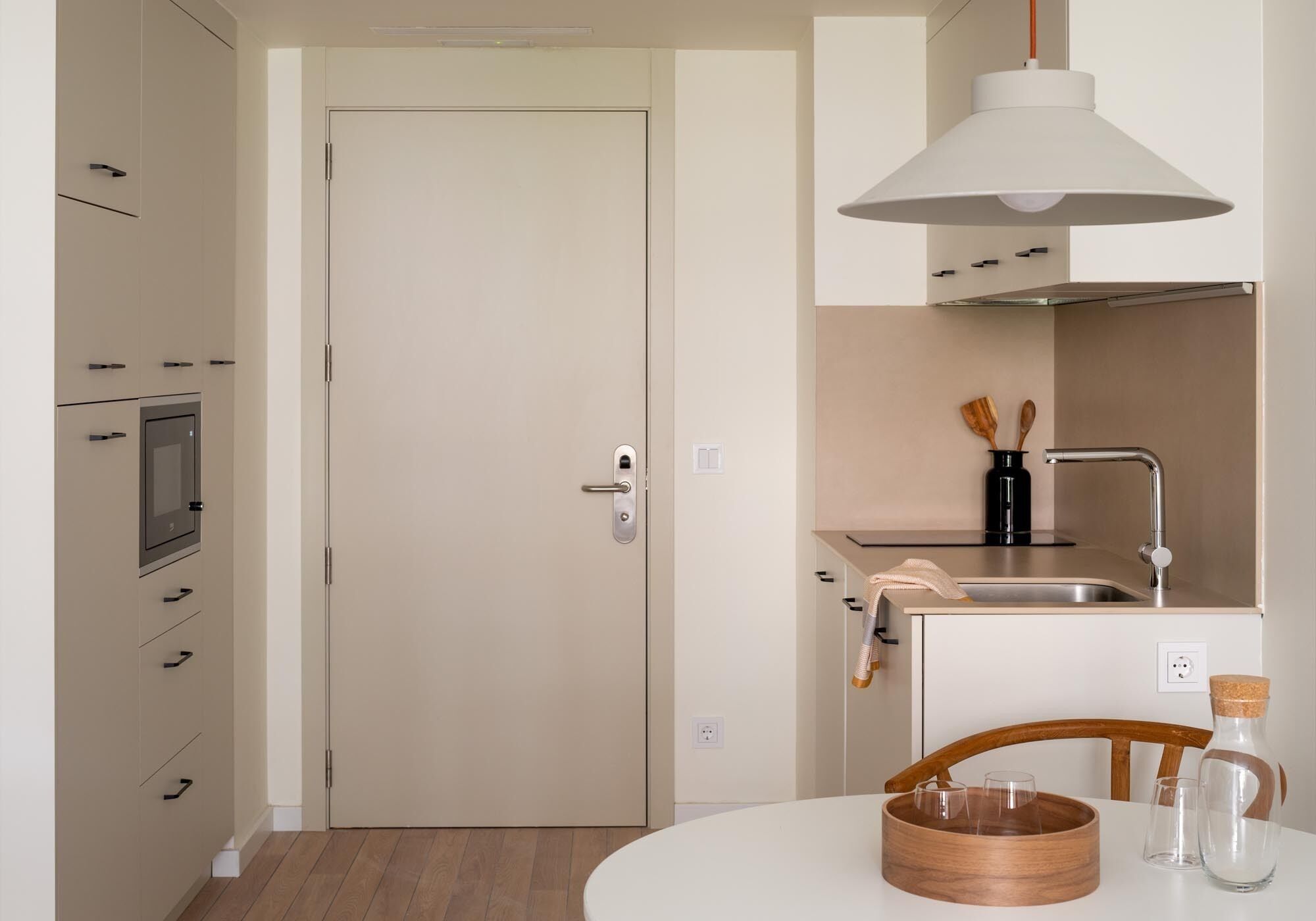 Una cocina moderna con puerta blanca, electrodomésticos integrados, un lavamanos y una mesa redonda de madera con vasos.