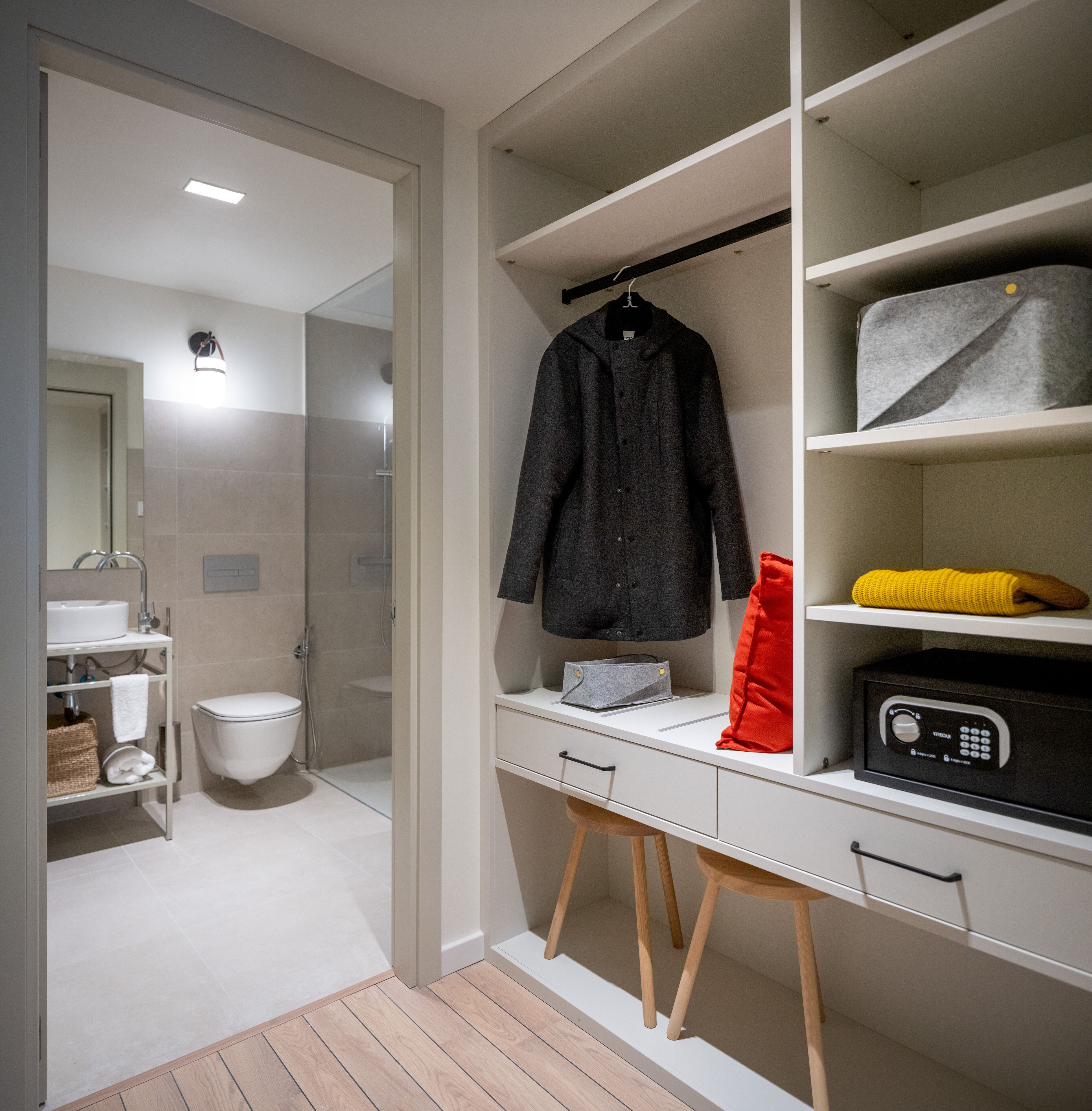 Un baño moderno con inodoro, lavabo y ducha. Se aprecia un tocador blanco con espejo, un perchero y una caja fuerte.