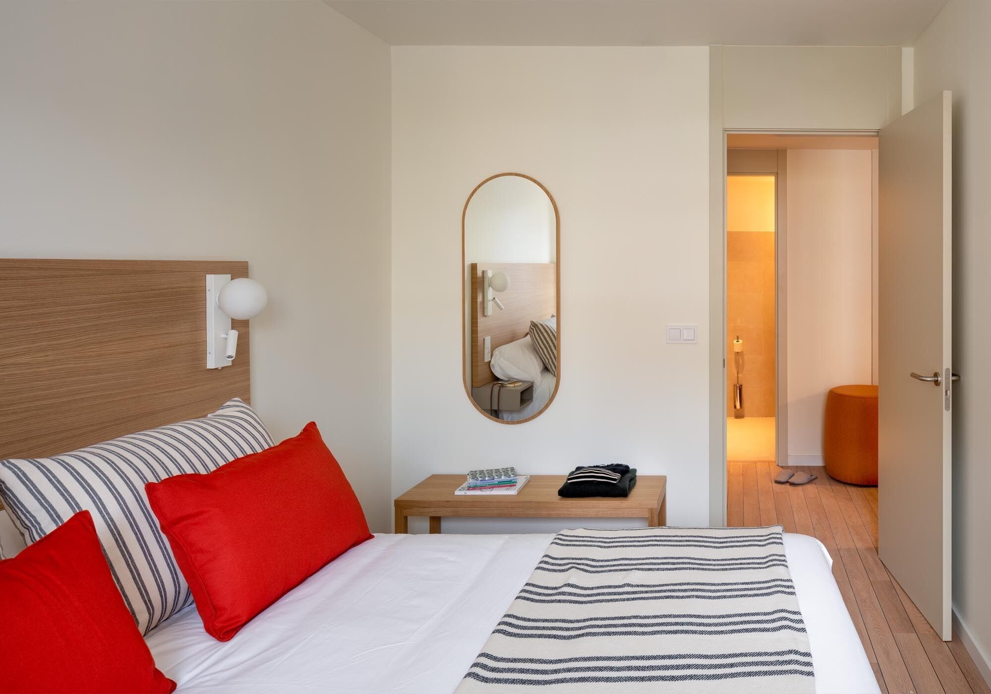 A hotel room with a bed, a bedside table with a lamp, a mirror, and a door leading to another room.