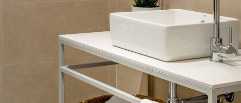 Un baño moderno con un lavabo blanco, grifo cromado y un espejo redondo. Hay una pequeña planta verde sobre el estante del lavabo y una cesta tejida debajo.