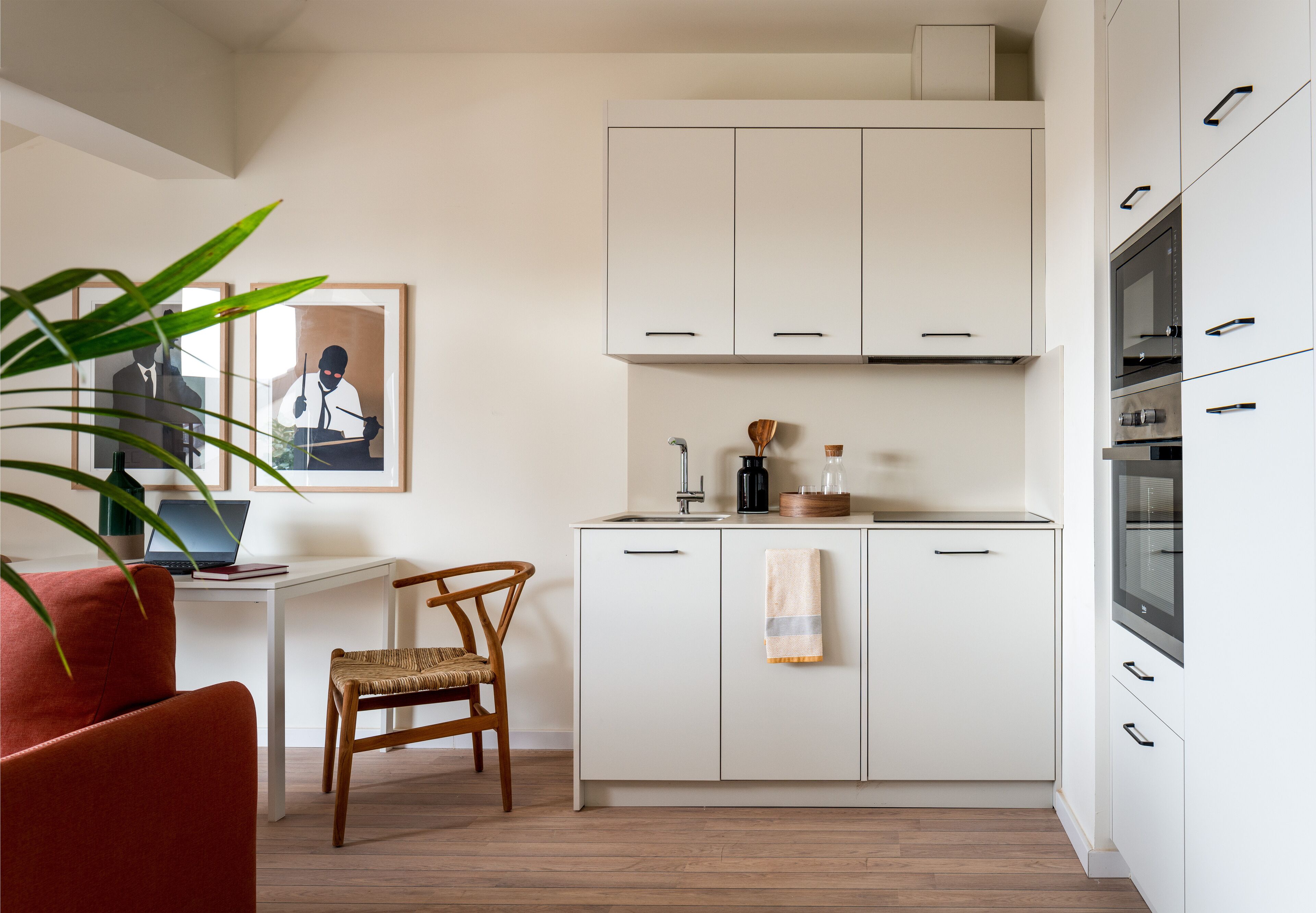 Une cuisine de taille réduite avec des meubles blancs, une chaise en bois, un ordinateur portable posé sur un bureau et un tableau encadré accroché au mur.