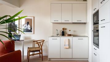 Una cocina compacta con armarios blancos, una silla de madera, una computadora portátil sobre un escritorio y un cuadro en la pared.