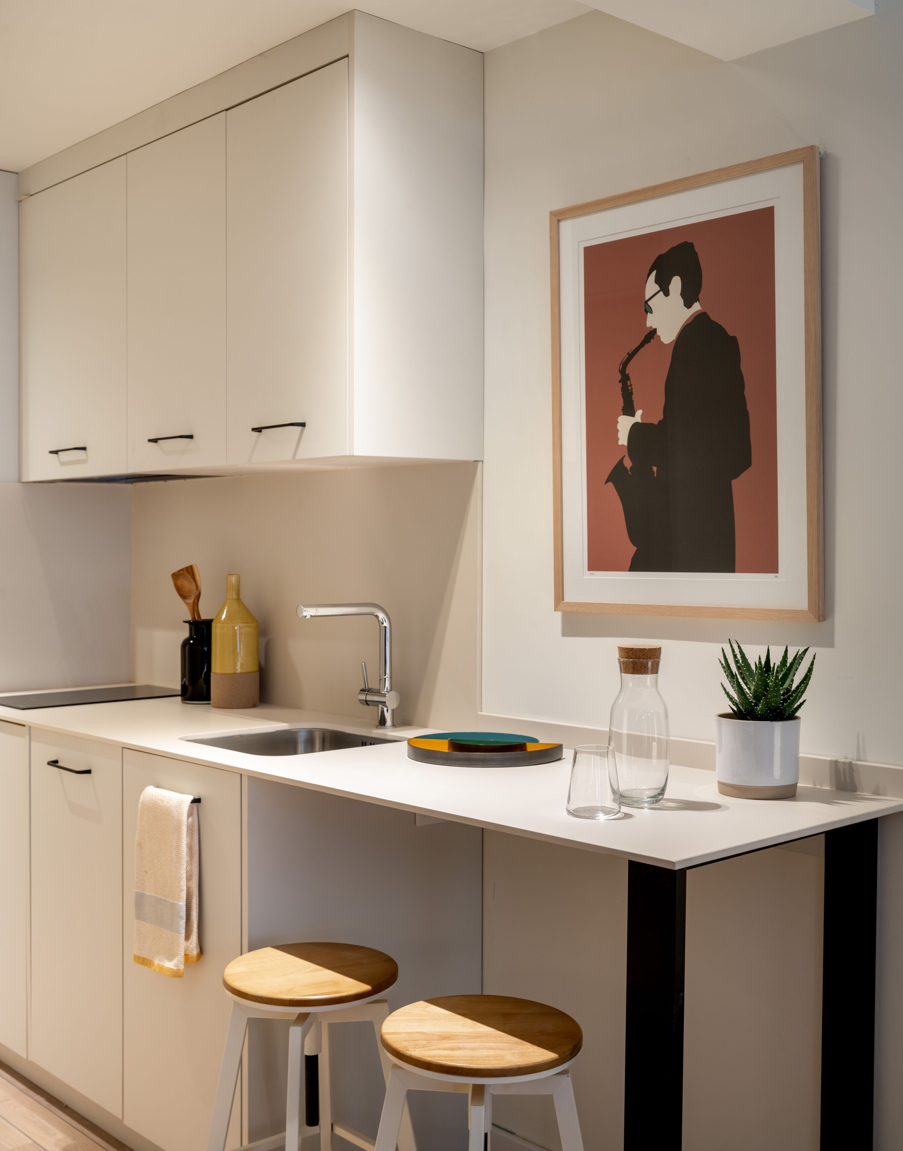 A modern kitchen with white cabinetry, a stainless steel sink, two wooden stools, and a framed artwork on the wall.