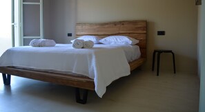 Un lit bien fait, avec du linge de lit blanc et des serviettes pliées, une tête de lit en bois et un petit tabouret noir à côté.