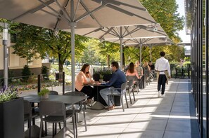 Un café en plein air avec des tables et des chaises sous des parasols, entouré de verdure et de bâtiments.