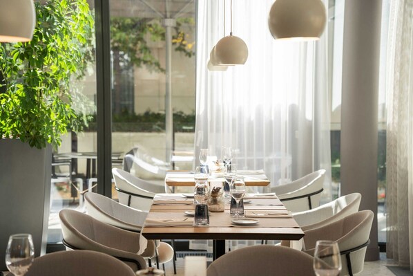A restaurant with a long table set for dining, large windows, and hanging pendant lights.
