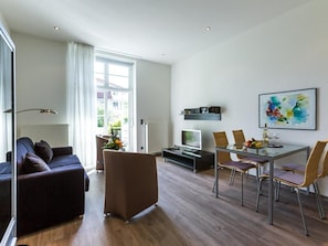 A modern living room with a sofa, a glass dining table with chairs, a flat-screen TV, and a painting on the wall.