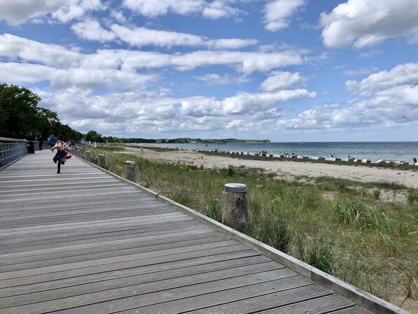 Ein hölzerner Steg führt zu einem Strand mit blauem Himmel und weiter Küste.