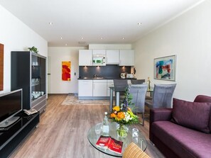 A modern living room with a glass coffee table, a purple sofa, and a kitchen in the background.