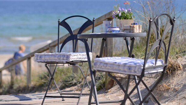 Una mesa junto a la playa con sillas, una mesita y una planta en maceta.