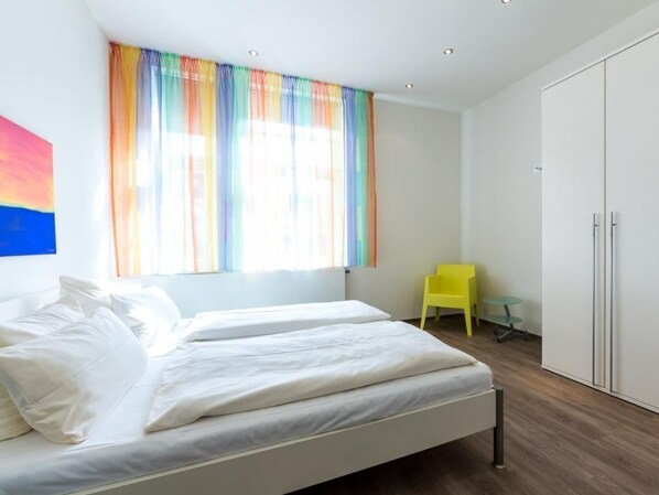 Une chambre avec deux lits, une fenêtre ornée de rideaux colorés, une chaise jaune et une armoire.