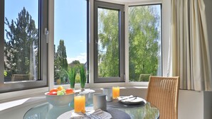 A dining area with a glass table, wicker chair, and a view of trees through large windows.