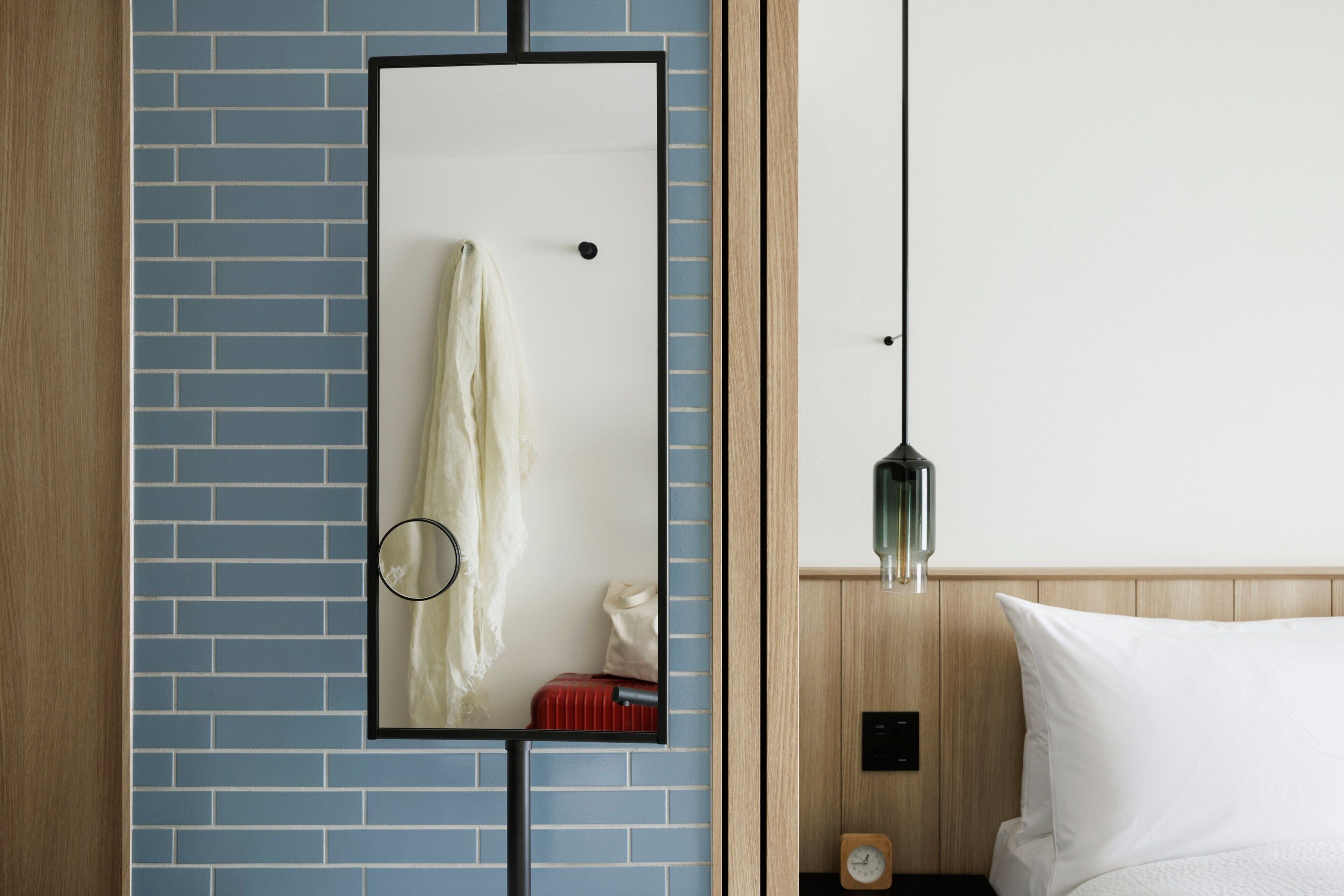 Une chambre d’hôtel avec un mur recouvert de carreaux bleus, une tête de lit en bois, un lit avec une literie blanche, une table de chevet avec une horloge et un miroir qui reflète un rideau de douche et une valise.