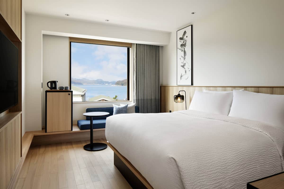 A modern hotel room with a large bed, a view of the sea, a small seating area, and a flat-screen TV.