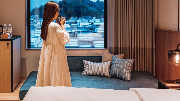 Ein Hotelzimmer mit großem Fenster, einem Bett, einer Couch und einem Schreibtisch mit Lampe.