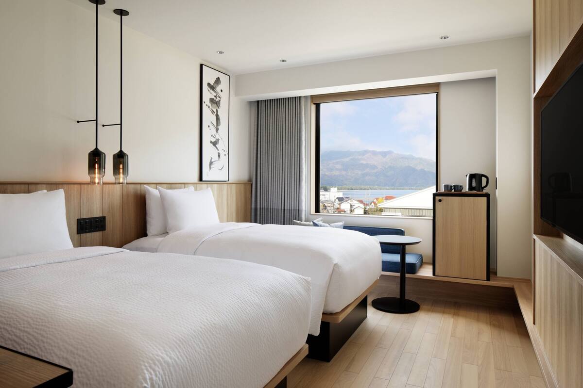 A hotel room with two beds, a large window with a view of mountains, a small table with a chair, and a TV on the wall.