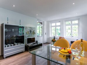 A modern living room with a glass dining table, a TV, and a bookshelf.